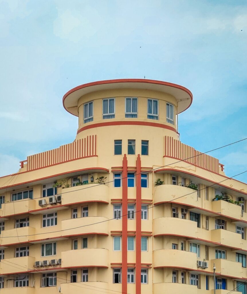 Yellow Art Deco–style apartment building with rounded balconies, vertical red accents, and a curved rooftop pavilion under a clear blue sky.
